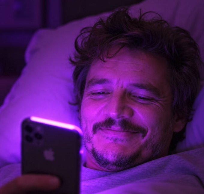 A man with curly hair lies in bed bathed in purple light, smiling contentedly as he looks at an iPhone.