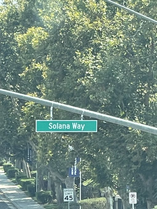A green street sign reads "Solana Way" hanging above a tree-lined road, also displaying a 45 mph speed limit sign nearby. The image playfully captures the crypto reference in the post, showing an actual street named after the popular blockchain.
