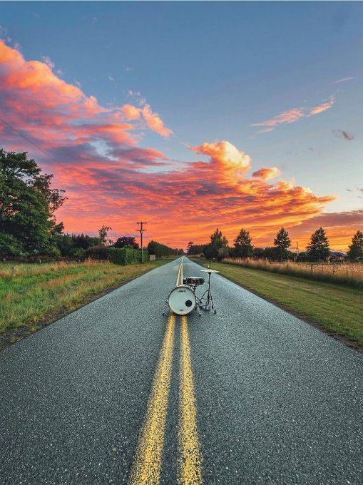 A scenic view of an empty two-lane road stretching toward the horizon under a dramatic sunset sky filled with pink and orange clouds, with a drum set placed in the middle of the road.