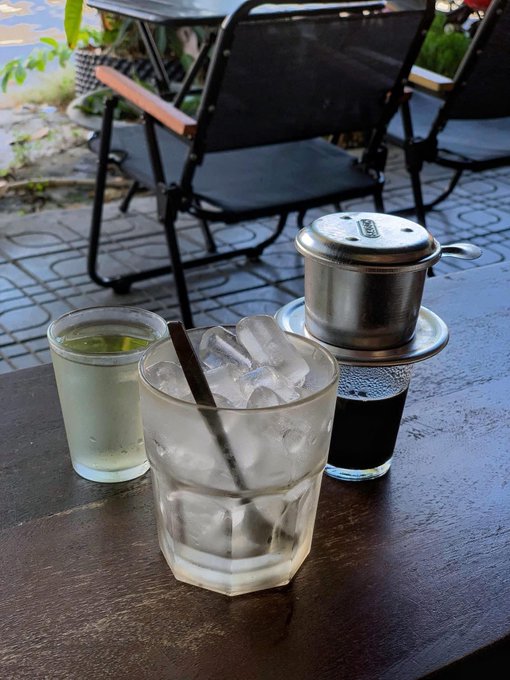Two glasses—one containing green tea and the other an iced drink with a straw—sit on a wooden café table next to a traditional Vietnamese phin coffee filter dripping dark coffee into a glass below, capturing a relaxing weekend morning coffee ritual.