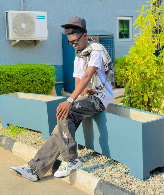 A young man in a white t-shirt, black pants, cap, sunglasses, and a patterned scarf sits on a blue planter box in an outdoor setting.