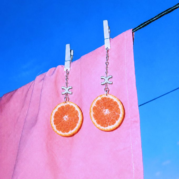 A pair of handmade earrings featuring dried orange slices hanging from clothespins against a pink cloth and blue sky.