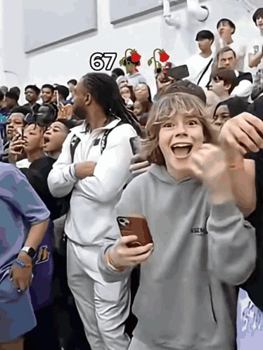 The image shows a crowd of people gathered together, with one person holding up their phone and showing off something on the screen. This individual appears to be excited or engaged with whatever they are viewing on their phone. The surrounding crowd seems interested as well, creating an atmosphere of shared curiosity and enjoyment.
