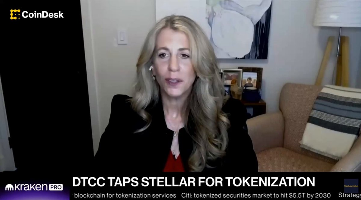 In the image, a woman with blonde hair is sitting in a chair while wearing a black jacket. She appears to be giving a presentation or speaking at an event. The background of the image features a painting hanging on the wall behind her. There are also some text elements visible in the image that read "@DenelleDixon" and "@CoinDesk".
