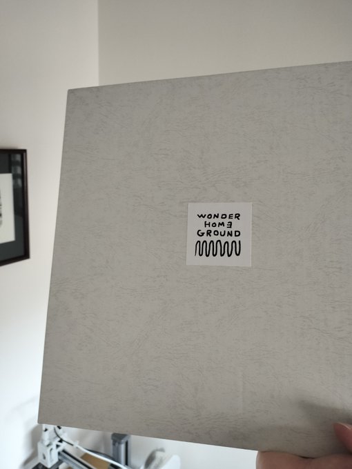 A person holding a textured light gray record sleeve or board with a small white sticker reading 'WONDER HOME G-ROUND' with a wavy line design, photographed in what appears to be a room with a framed picture on the wall.