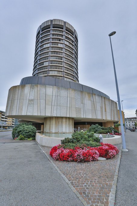 A photograph of a distinctive circular building with a tall cylindrical tower, surrounded by a plaza with red flowers and a street lamp.