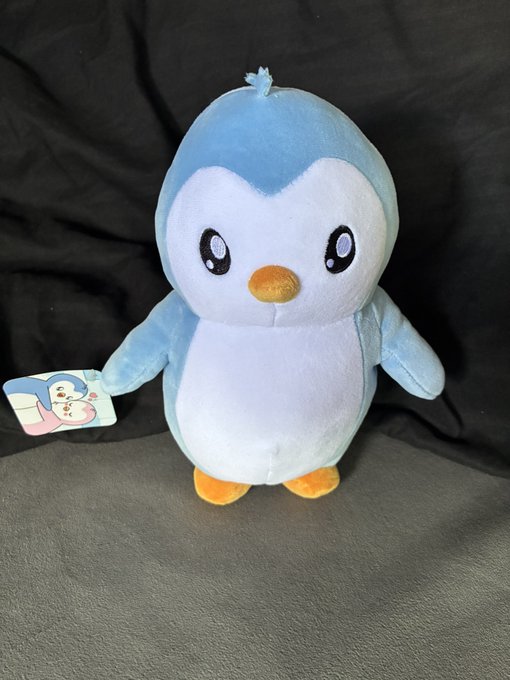 A blue and white plush penguin toy with large sparkly eyes and an orange beak sits against a black fabric background, with a tag attached to its side.