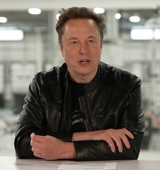 Elon Musk sits at a table in a black leather jacket, speaking with his arms crossed in a bright indoor setting. The image accompanies a post joking that “Sir Doge” raises his SpaceX price target 69 times to 69,420,420 “Space Bucks,” referencing Dogecoin and Musk’s association with it.
