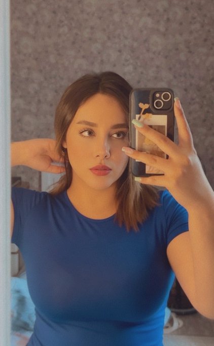 A young woman with shoulder-length brown hair in a blue fitted t-shirt taking a mirror selfie with her phone, which has a butterfly design on the case.