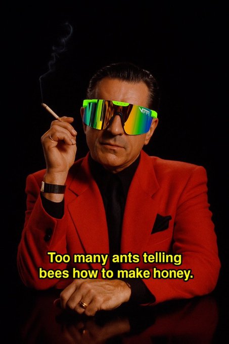 A man wearing a red suit, glasses, and sunglasses sits at a desk with a table lamp and cigarette behind him. The caption above him reads "Too many ants telling bees how to make honey." It seems to be referencing an article about something related to money or investing that was posted on the same website as this image.