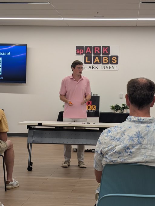 A man in a pink polo shirt presents in a room with a 'spark LABS by ARK INVEST' banner on the wall, with a TV screen and seated audience members visible.