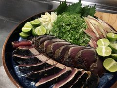 A photograph of a plate containing sliced raw fish, likely tuna sashimi, garnished with lime wedges, chopped green onions, and other accompaniments.