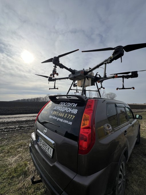 A drone with multiple rotors is mounted on the roof of a black SUV that has Ukrainian text 'поспіти АГРОДРОНІВ' and a phone number on its rear window.