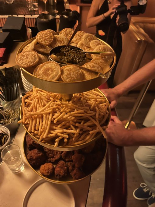 A three-tiered gold serving tray holding bread with caviar, french fries, and fried balls, with a photographer holding a camera visible in the background.