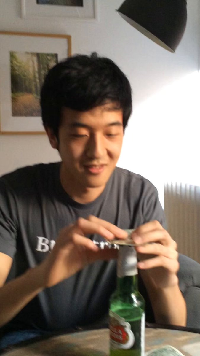 The image features a man sitting at a table with a green beer bottle, possibly opening or closing the cap. The man appears to be smiling as he engages with the beer bottle. In the background, there are two pictures hanging on the wall above the man's head and another picture in front of him.