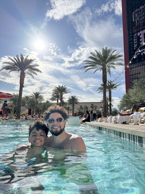 A man with sunglasses and a young boy smile together in a swimming pool with palm trees and a sunny sky in the background.