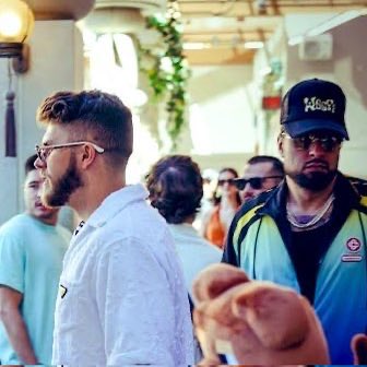 A man wearing a black 'Money Mike' cap, sunglasses, and a turquoise/black shirt stands next to a bearded man in glasses and a white shirt, with another person in the foreground.