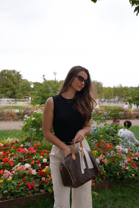A woman with long brown hair and sunglasses stands outdoors in a park holding a Louis Vuitton Speedy handbag, with colorful flower beds and trees in the background.