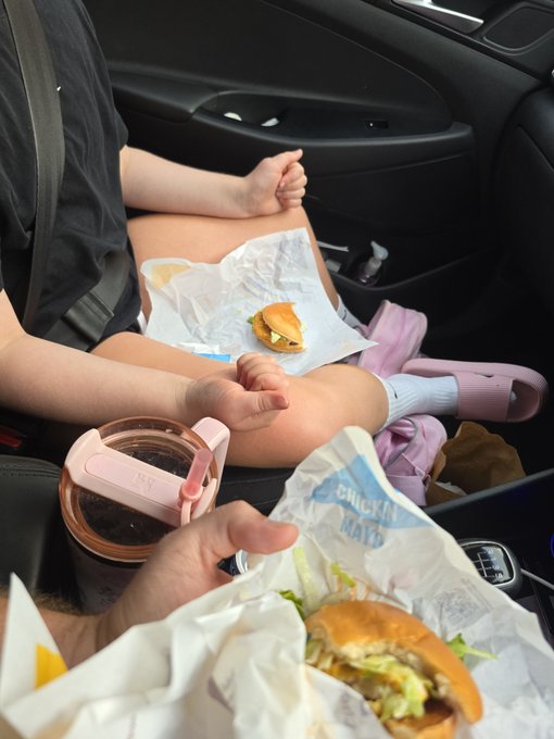 Two people eating Chick-fil-A sandwiches and drinking from a tumbler while sitting in a car.