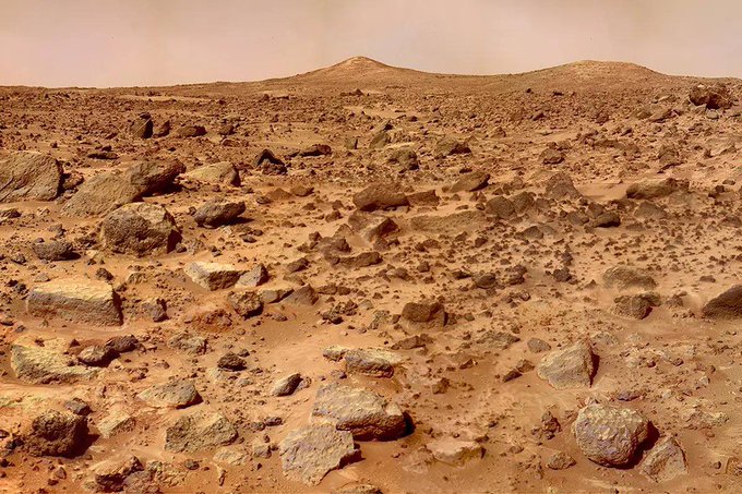A photograph of the Mars surface showing a rocky, barren reddish-brown desert landscape with scattered boulders and distant hills.
