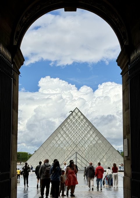 The image shows a large group of people walking around outside an archway, passing through a doorway that leads to the Louvre Museum in Paris, France. The sky above them is filled with clouds, creating a dramatic backdrop for this scene. In the background, there are several people carrying backpacks and handbags, possibly tourists exploring the city or visiting the museum. The image also contains text that reads "Cosmos has joined the @UNDP Blockchain Advisory Group (BAG), a senior-level advisory forum organized by the United Nations Development Programme." This suggests that the group of people might be tourists who have just arrived at the Louvre and are now walking towards the entrance, possibly discussing their plans for exploring the museum.