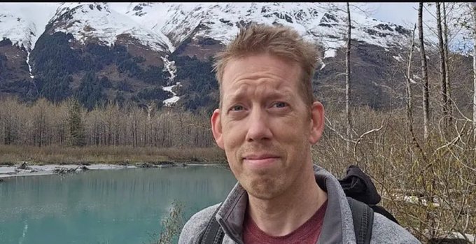 A man with reddish hair takes a selfie in front of a turquoise river with snow-capped mountains and bare trees in the background.