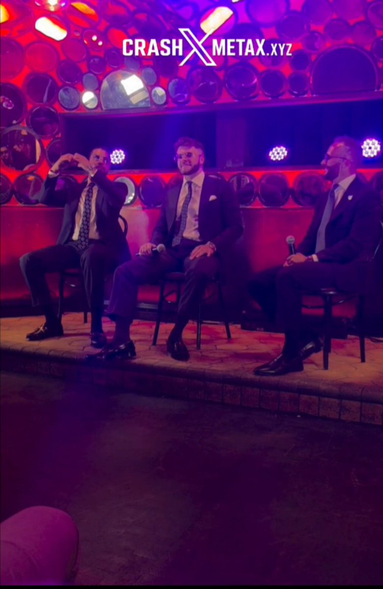 Three men in suits sit on chairs holding microphones on a red-lit stage with a 'CRASH METAX.XYZ' logo displayed above them.