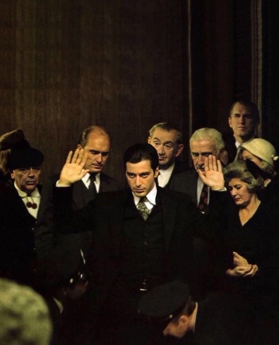 A cinematic scene of a young man in a dark suit raising his hands among a crowd of formally dressed people in a dimly lit setting, from what appears to be The Godfather film.