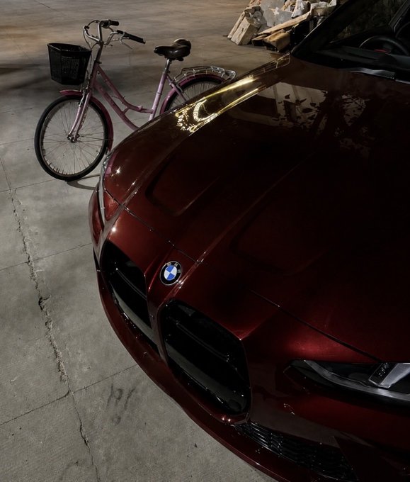 A dark red BMW with glossy paint is parked in a concrete garage next to a pink bicycle with a front basket.