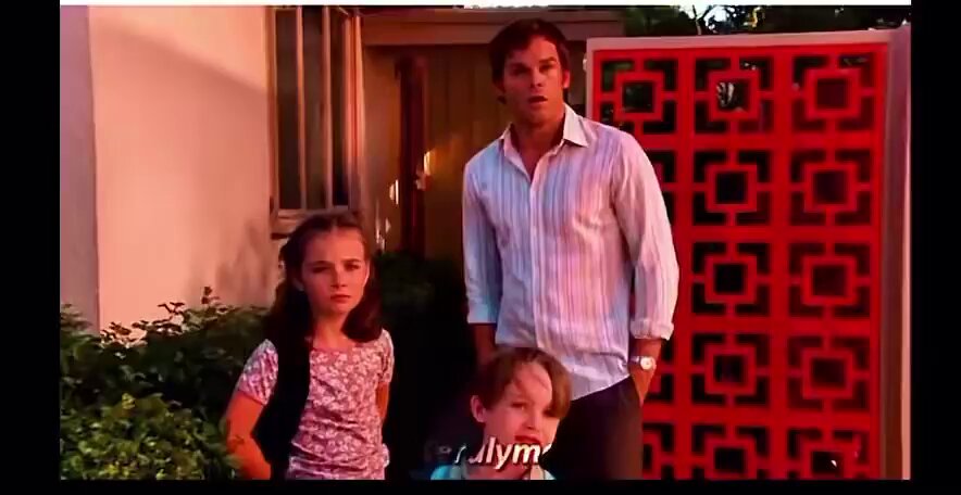 The image shows a man standing next to two children outside of a house, with a red door behind them. The scene appears to be set at dusk or dawn when the lighting is soft and warm. There are no texts or other objects visible in the image, but there is a clock on one wall that reads "9:00".