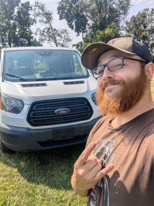 A man wearing glasses, a baseball cap, and a graphic t-shirt flashes a peace sign in front of his white Ford work van parked on grass, celebrating a successful week with a raise, overtime pay, and a new company vehicle in this Day 607 self-portrait.
