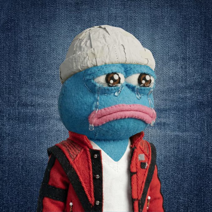A sad-looking blue plush toy character with a white cap, large eyes, pink lips, and embroidered tears wears a red jacket over a white shirt against a denim background.