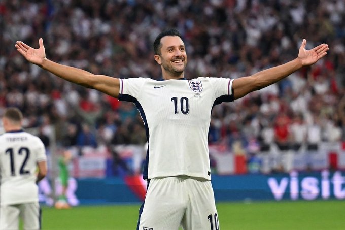 A soccer player in a white England jersey with the number 10 celebrating with arms outstretched on the field, with another player wearing number 12 visible in the background.