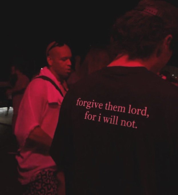 Photo taken at a dimly lit event with red lighting, featuring a person wearing a black shirt that reads 'forgive them lord, for i will not.'