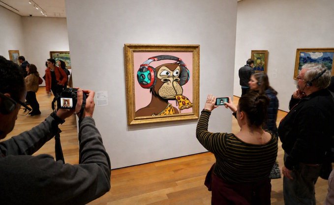People in an art gallery photographing a colorful, cartoon-style painting of an owl wearing headphones in an ornate gold frame.