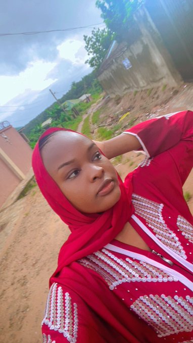 A selfie of a woman wearing a red headscarf and a red beaded dress, taken outdoors with a dirt path and buildings in the background.