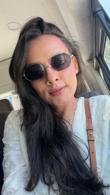 A selfie of a woman with long dark hair wearing dark sunglasses and a white top, seated inside a vehicle.