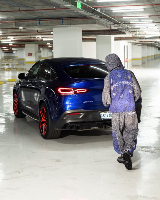 A person wearing a blue star-patterned 'goooaaaal' hoodie walks toward a blue Mercedes GLE Coupe with red accents in an underground parking garage.