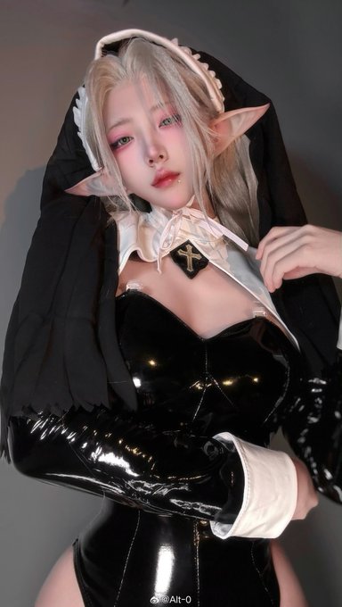 A person with pointed elf ears, blonde hair, and dramatic makeup poses in a black latex nun costume with a white veil featuring a cross emblem. The watermark "Alt-0" appears in the bottom right corner, identifying the cosplayer credited in the accompanying post.