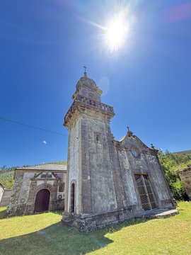 An old stone church with a tall bell tower stands under a bright, sunlit sky in San Xurxo de Moeche, Galicia. The 11th-century building features weathered stone walls, a small cross atop the tower, and an arched wooden doorway, reflecting the historic heritage of the grandmother's family connection to this ancestral site.