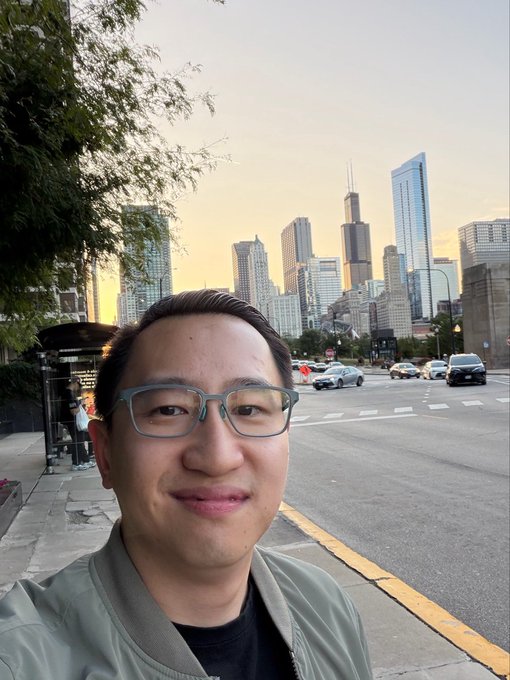 A smiling man wearing glasses takes a selfie on a city street with the Chicago skyline, including the Willis Tower, visible in the background at sunset.
