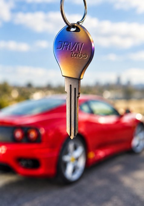 A shiny metallic key with a colorful head engraved with 'DRVN labo' hangs in front of a blurred red sports car parked under a partly cloudy sky.