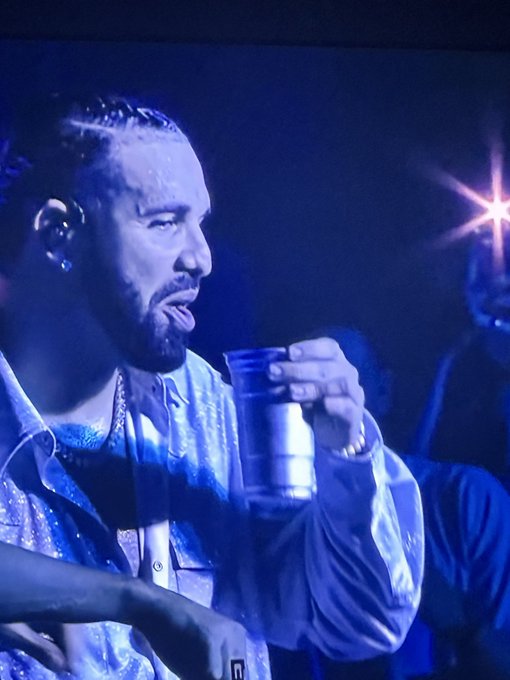 A man with braids and a beard performing on stage under blue lighting, holding a cup while speaking or rapping into a microphone.