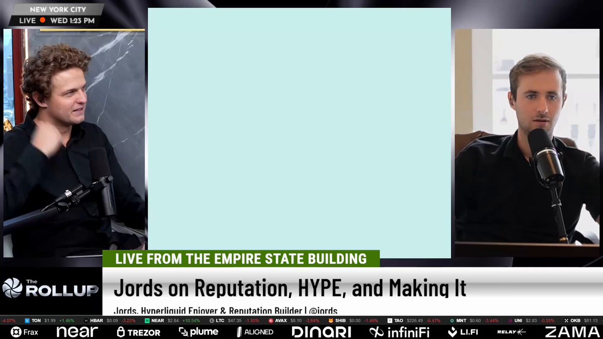 The image shows a television screen with two men sitting behind microphones, appearing to be discussing something related to the Empire State Building. One man is wearing a black shirt while the other has a blue shirt. They are both talking into their respective microphones, which can be seen clearly on the screen. The background of the image features a white square that reads "Live from the Empire State Building".