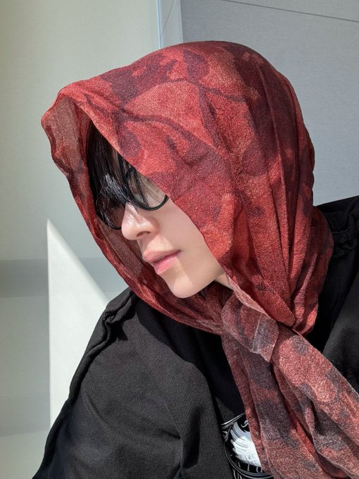 A close-up selfie of a person with glasses wearing a sheer red floral scarf draped over their head.