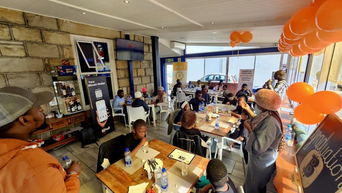 A crowded indoor gathering, possibly a community event or 'Bamba Bula Walla' meeting, featuring a 'Y' logo sign and many people seated at tables with orange balloons decorating the venue.