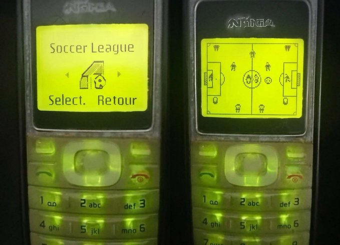 Two Nokia mobile phones display the classic "Soccer League" game menu and top-down field view on their yellow backlit screens, evoking nostalgic memories of early 2000s mobile gaming. The French word "Retour" (Return) is visible on the left screen's menu, and the iconic numeric keypad glows beneath each display.