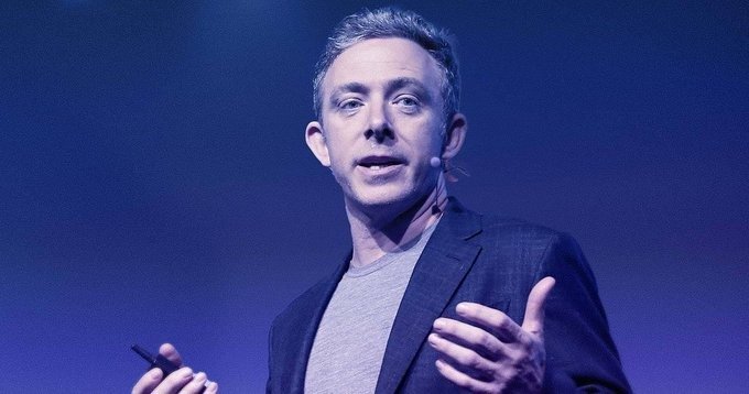 A man with short hair, wearing a dark blazer over a gray t-shirt and a headset microphone, speaks expressively on a stage bathed in blue lighting while holding a small clicker. The image likely depicts a tech executive or speaker giving a presentation, visually complementing the post's discussion of $ICP's (Internet Computer Protocol) outdated all-time high and its current network development metrics.