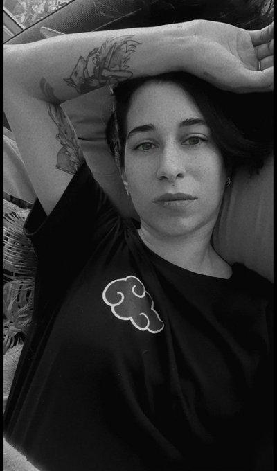 A black-and-white selfie of a person with short dark hair resting their tattooed arm on their forehead, wearing a black t-shirt featuring a cloud-like logo.