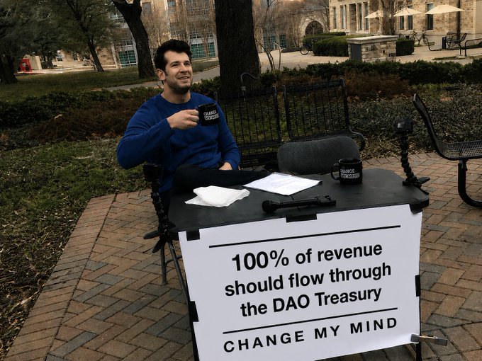 A man sits at an outdoor table holding a "Change My Mind" coffee mug, with a "Change My Mind" sign reading "100% of revenue should flow through the DAO Treasury," visually personifying the argument from the Ownership Coin Monthly report that all project revenue should first pass through the DAO treasury before being reinvested.